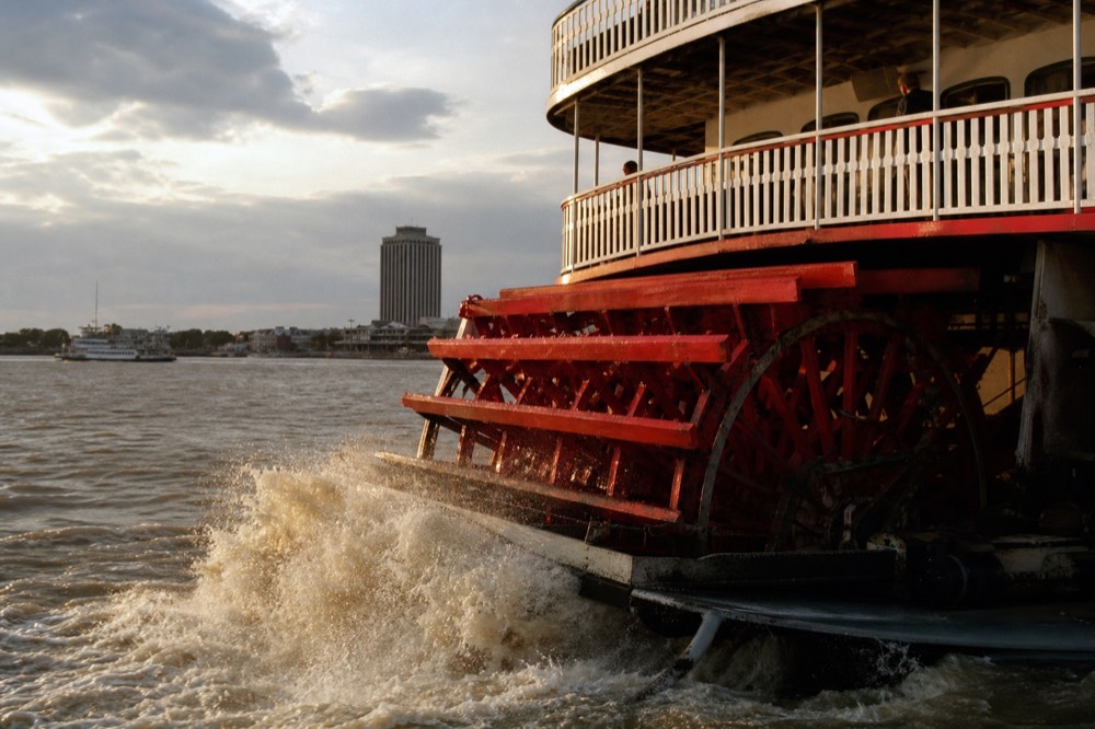 Red paddlewheel of a steam-powered sternwheeler churning brown Mississippi River water, white deck railings above it