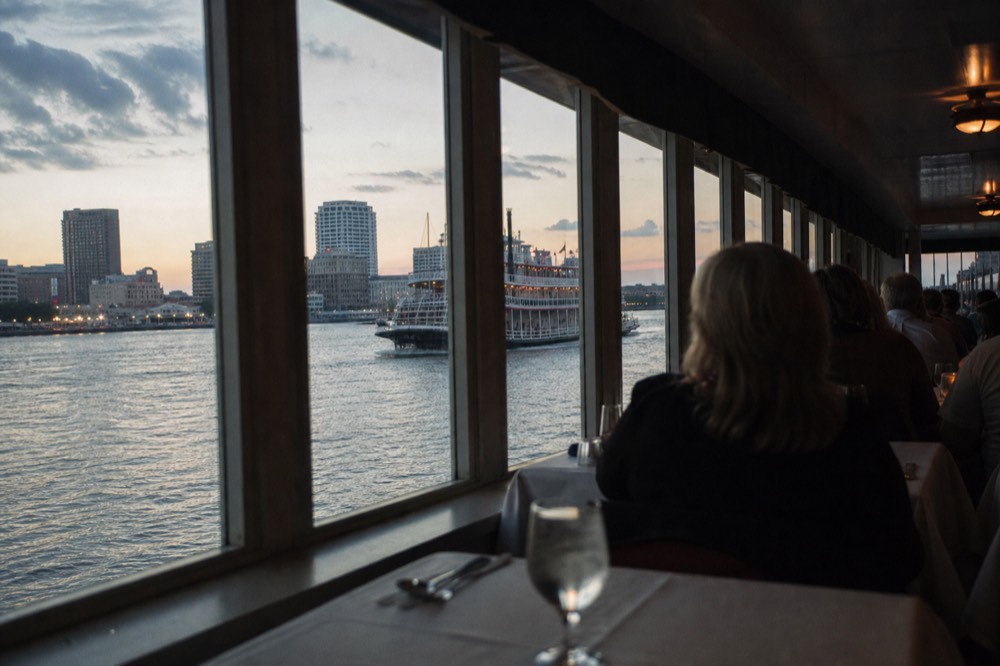Passengers seated at white-clothed tables in a riverboat dining room with tall windows showing the passing riverbank