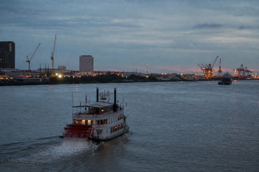 Wide view of the Mississippi crescent bend at New Orleans with a small excursion paddlewheeler in the foreground and port cranes downriver