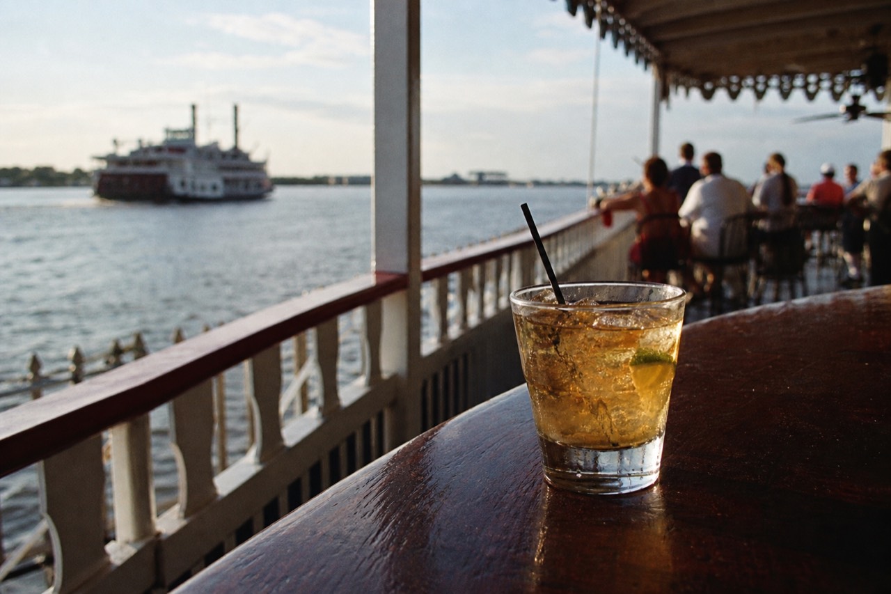 Cocktail glass on a riverboat bar top with the open deck and the Mississippi blurred behind it in evening light