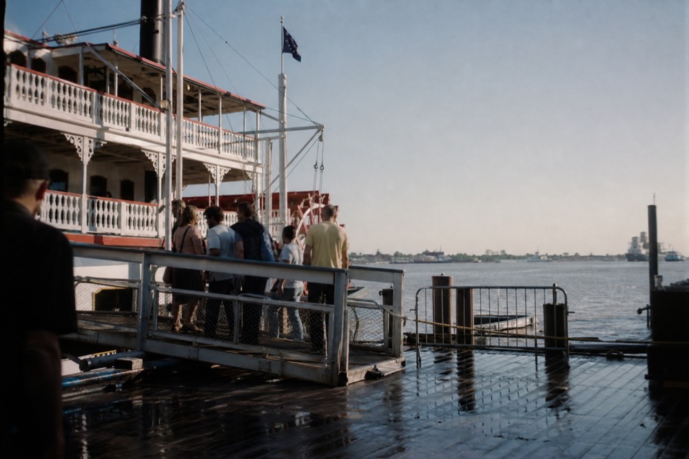Boarding gangway of a paddlewheeler at a riverfront plaza dock with passengers in light casual clothes stepping aboard