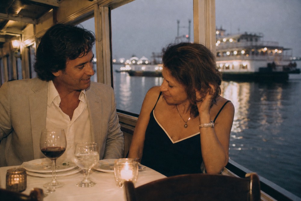 Couple in smart-casual evening clothes at a white-clothed table on a riverboat with warm lamplight and dark water outside the window