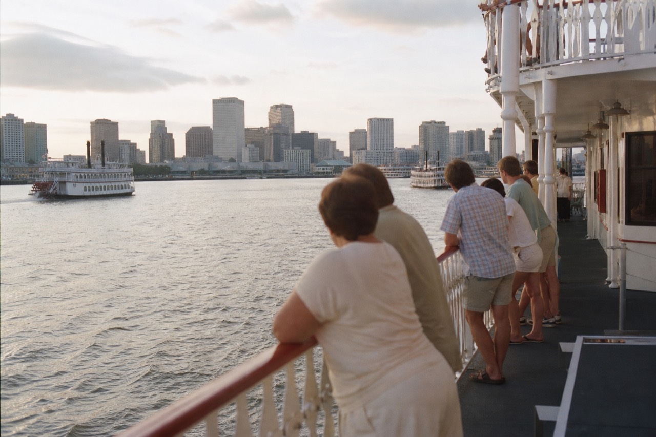 Passengers in light summer clothes standing along the rail of a white paddlewheeler at dusk with the New Orleans skyline behind