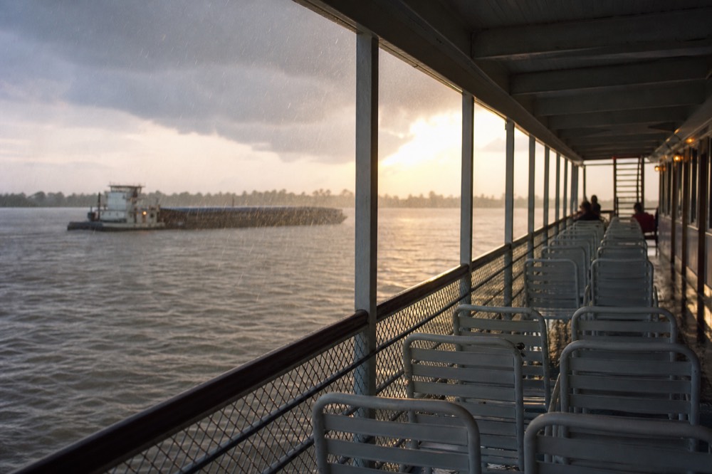 Covered riverboat deck with rows of empty white chairs under an overhang, rain streaking past the open side and the grey river beyond
