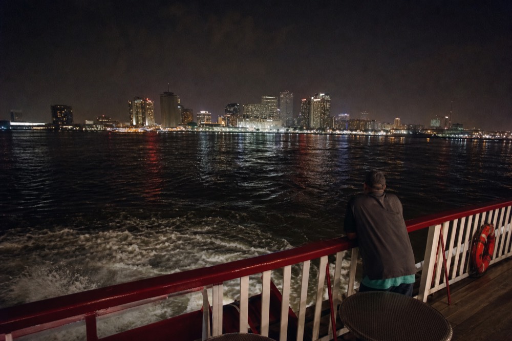 New Orleans skyline lit at night seen from the open rear deck of a riverboat with the paddlewheel wash below