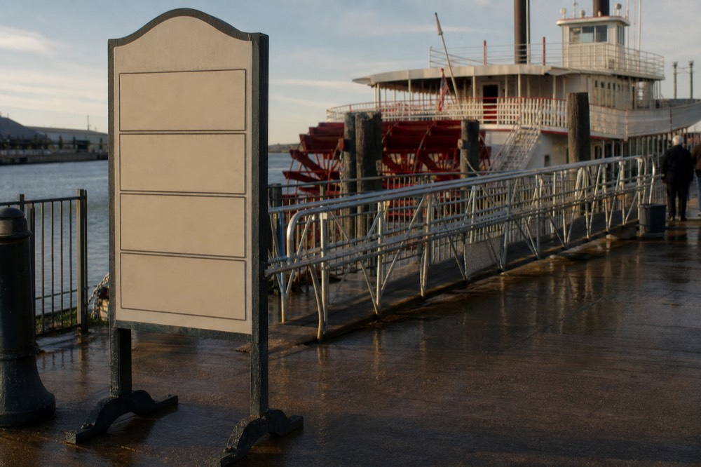 Fare board beside a riverboat gangway listing cruise times with the Mississippi and a moored sternwheeler behind