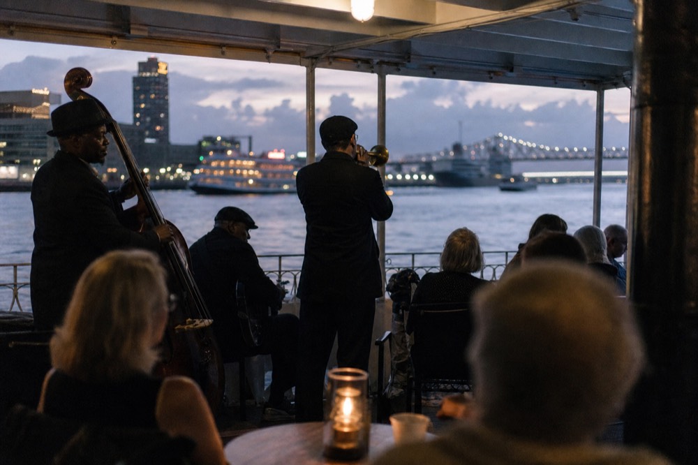 Jazz band playing on a riverboat deck in the evening with passengers seated close and the city lights beyond the rail