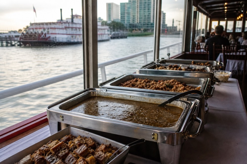 Creole buffet on a riverboat dining deck with gumbo, jambalaya and bread pudding in chafing dishes beside a window over the river