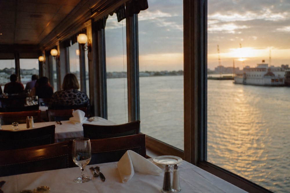 Dining room aboard a riverboat with white-clothed tables set for dinner and tall windows onto the dark river
