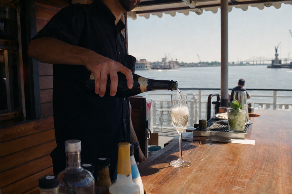 Bartender pouring a sparkling cocktail at a small wood-panelled bar on a riverboat deck with the river behind