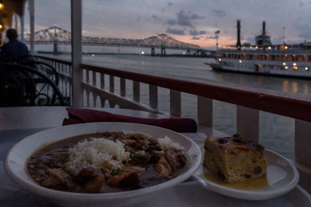 A bowl of dark seafood gumbo over rice and a square of bread pudding on a white plate at a riverboat table