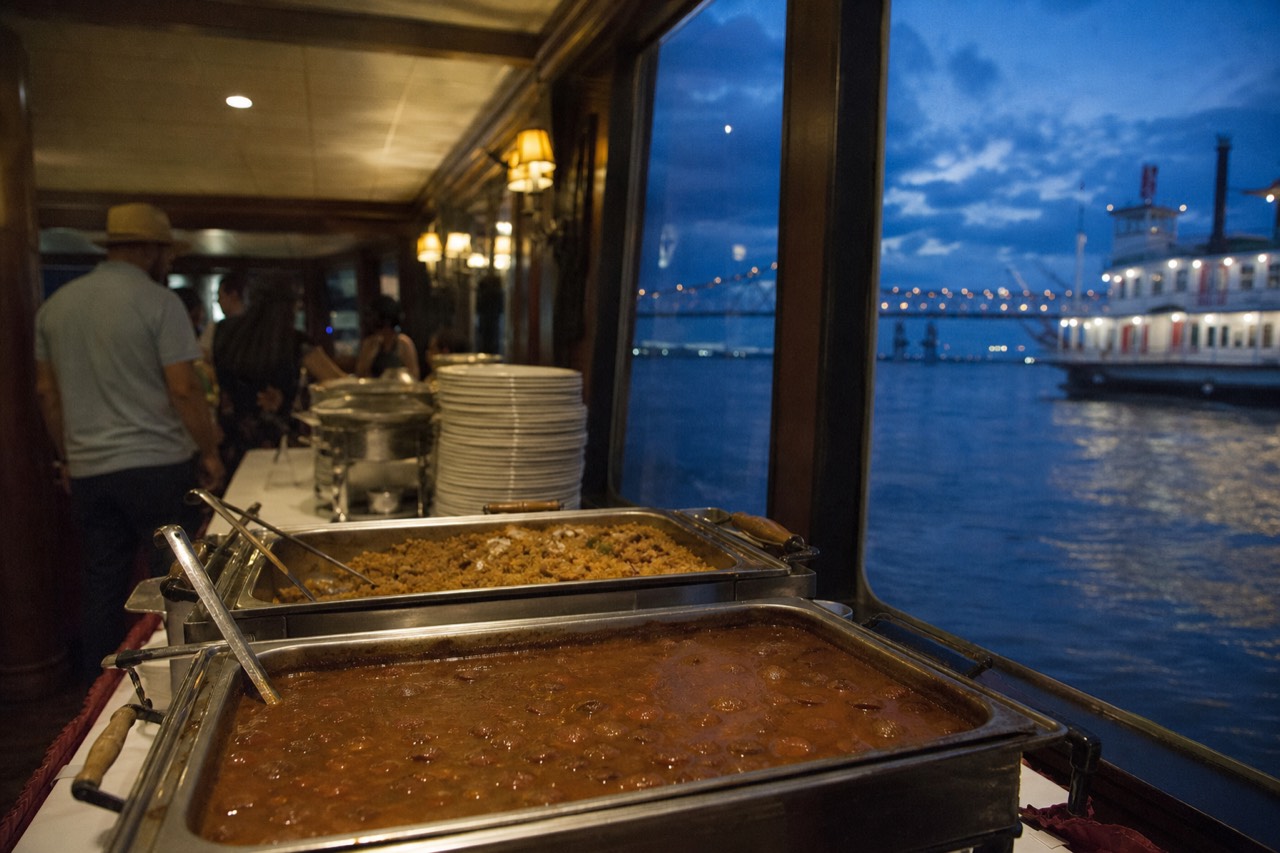 Creole buffet line aboard a riverboat with steam trays of jambalaya and gumbo and a stack of white plates