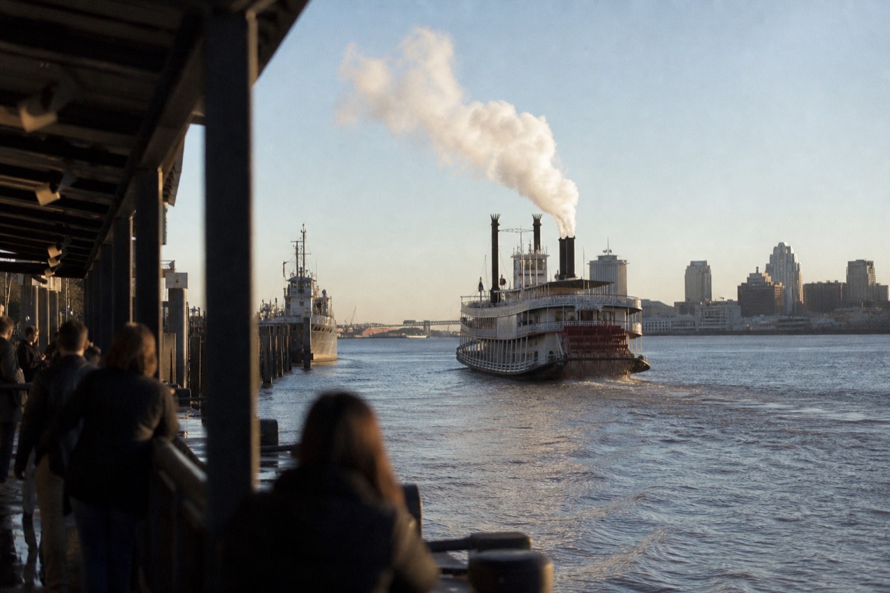 Steam sternwheeler with two tall black stacks pulling away from a New Orleans wharf with white steam rising from her whistle