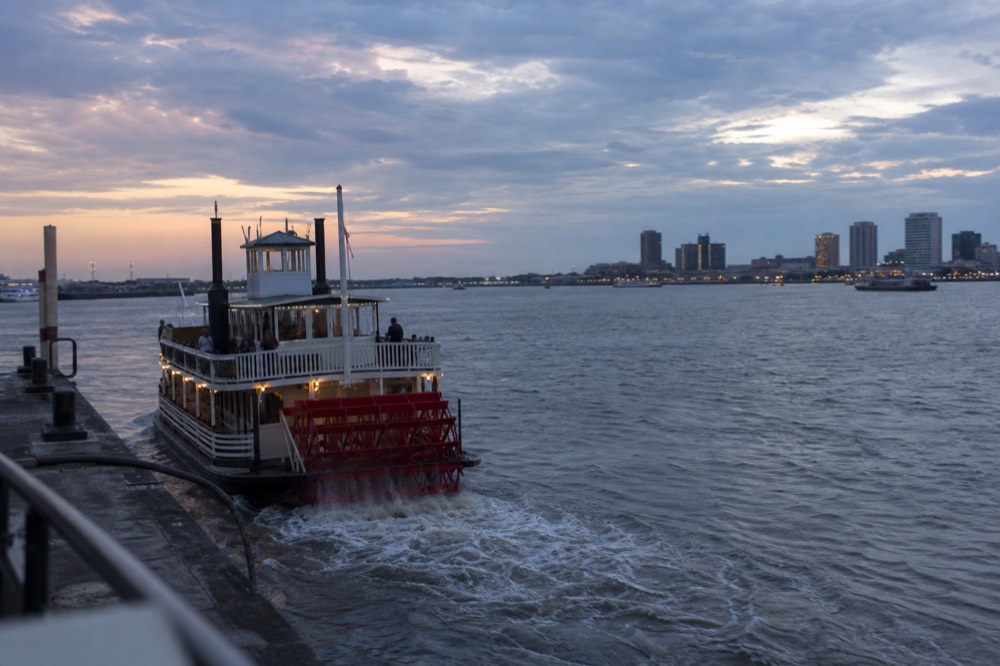 Smaller modern riverboat with a red paddlewheel departing a New Orleans wharf in late afternoon light