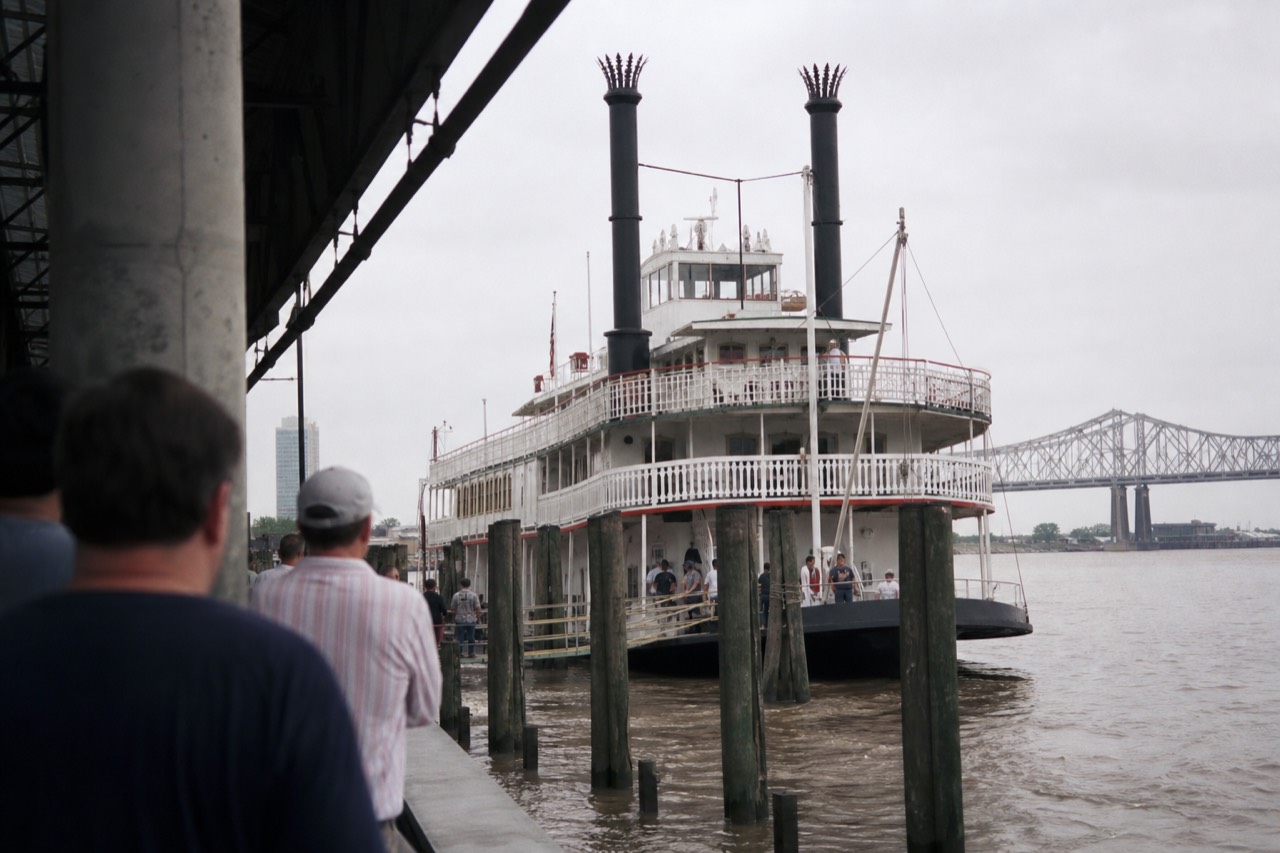 White steam sternwheeler with tall black stacks tied up at a New Orleans wharf while passengers wait on the levee to board
