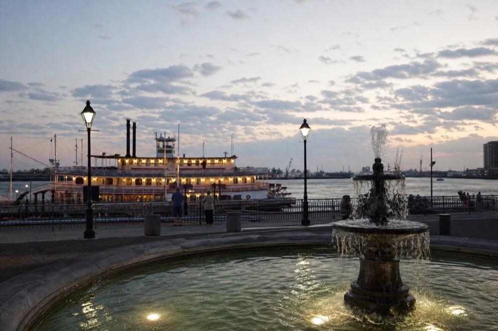 Evening view of Spanish Plaza in New Orleans with the fountain in the foreground and a lit paddlewheel riverboat moored at the dock beyond