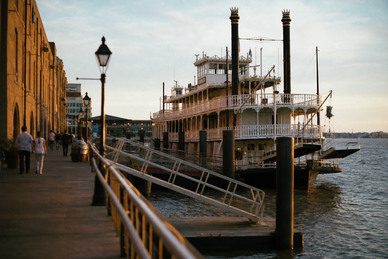 A riverfront wharf gangway in New Orleans with a white paddlewheel riverboat moored alongside and the brick facade of a French Quarter warehouse behind