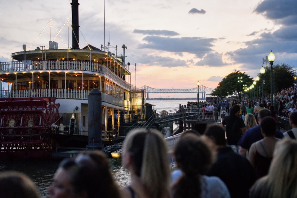 Crowded riverfront promenade at dusk during a festival weekend with a lit paddlewheeler at the wharf