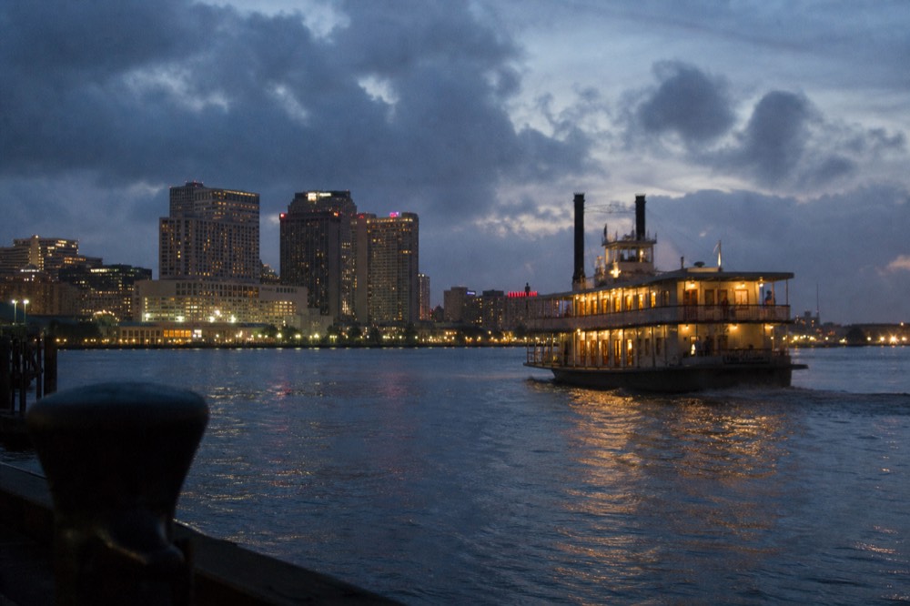 Riverboat pulling away from the wharf at dusk with the New Orleans skyline lit behind and warm light in the dining room windows