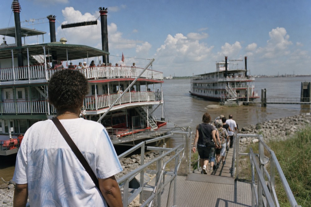 Passengers walking down a metal gangway from a riverboat onto a grassy levee path under a wide flat sky