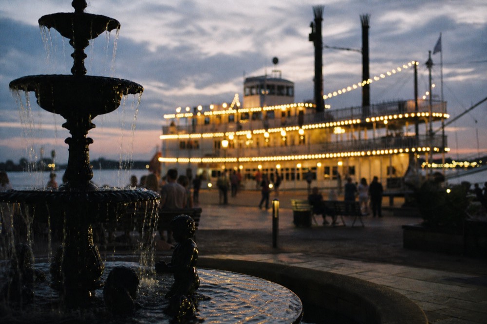Riverfront plaza at dusk with a moored paddlewheeler lit along its rails and a fountain in the foreground