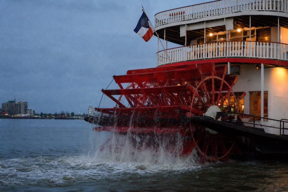 Red paddlewheel turning at the stern of a white riverboat, water streaming from the buckets