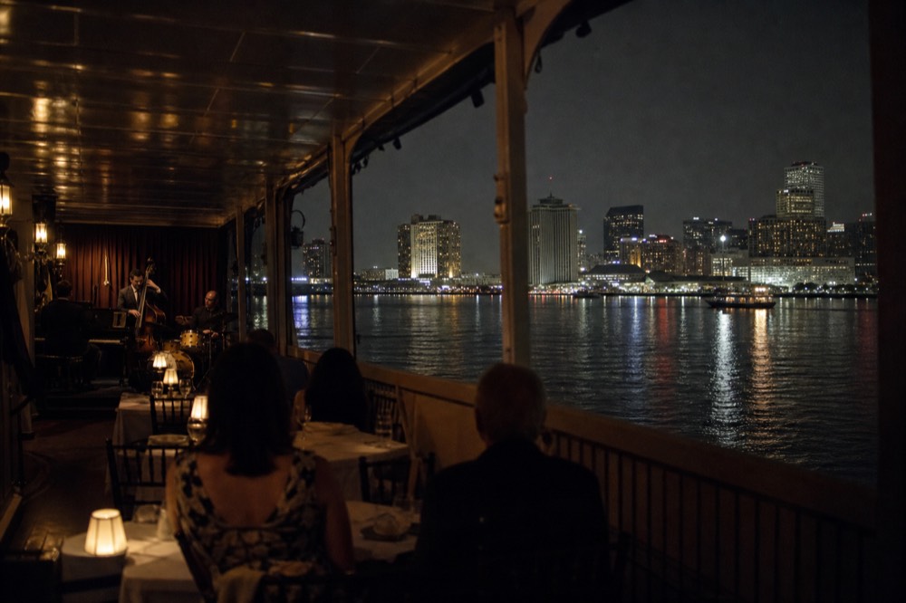 Riverboat dining room at night with white tablecloths, warm lamps and a jazz trio playing at the far end