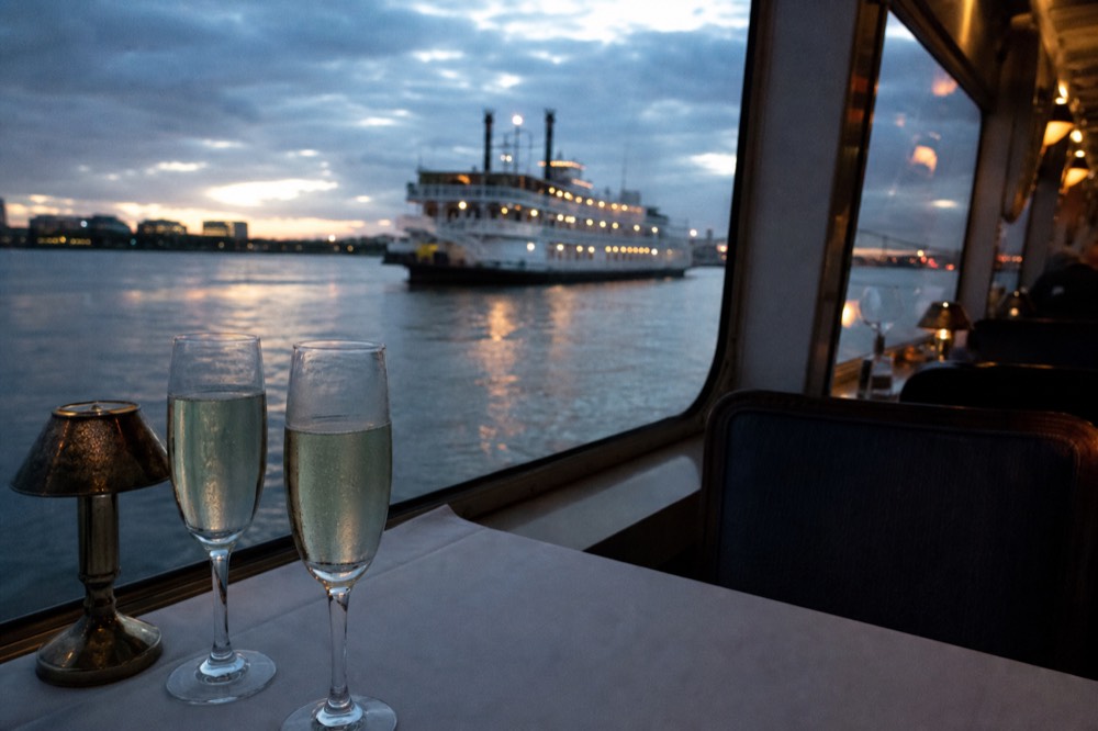 Champagne flutes on a white-clothed table beside a riverboat window with dusk light on the water outside