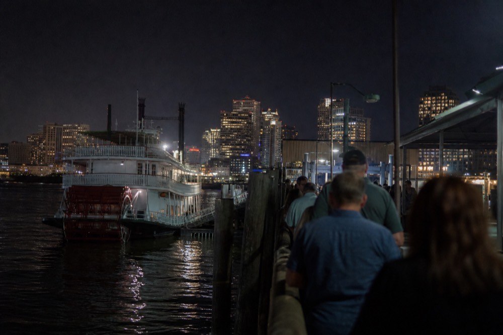Passengers waiting in a boarding line on a wooden wharf beside a moored white sternwheeler, morning sun on the water