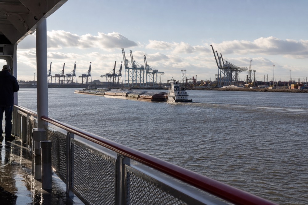 View from a riverboat deck of container cranes and a barge tow on the Mississippi under bright late-morning light