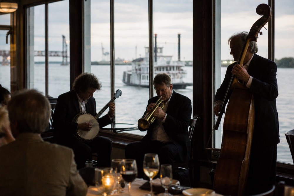 A small traditional jazz trio playing trumpet, banjo and upright bass in a riverboat dining room with tall windows onto the river
