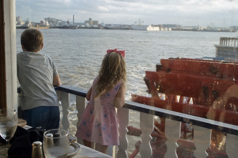Children leaning on a riverboat deck rail watching a red paddlewheel churn brown river water