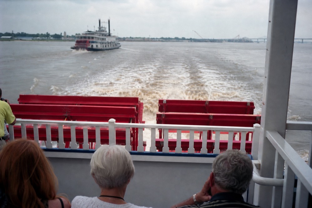 Passengers on the open upper deck of a sternwheeler in afternoon light with the paddlewheel churning brown river water behind