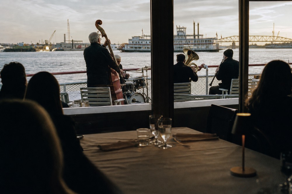 View from a dining room table toward a jazz band on a riverboat, with river water visible through the window beyond