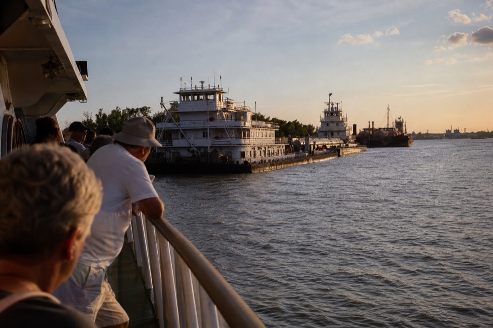 Passengers lining a riverboat upper deck rail in afternoon sun as the boat passes moored barges