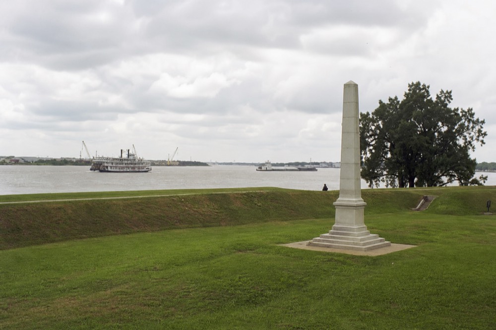 Green battlefield lawn with a tall white marble monument and a low earthen rampart under a wide Louisiana sky