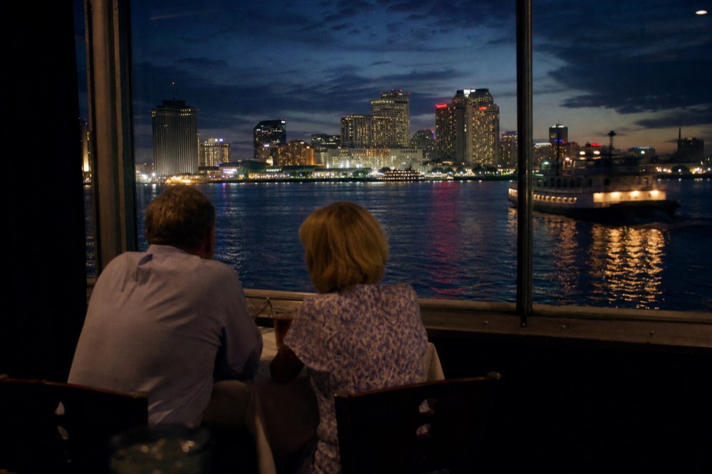 Couple seated at a riverboat window table at dusk with the lit New Orleans skyline reflected on the water