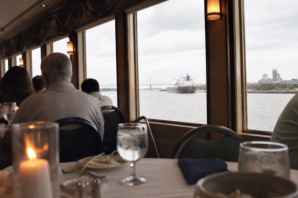 Passengers seated at white-clothed tables in an air-conditioned riverboat dining room with tall windows facing the river
