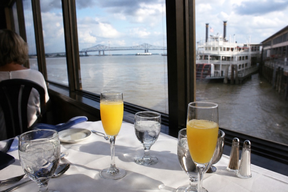 Champagne flutes of mimosa on a white tablecloth beside a riverboat window with the levee visible outside
