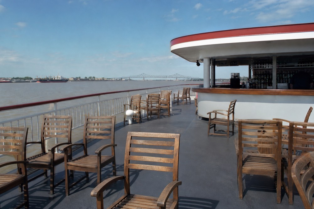 Riverboat rooftop deck with empty wooden chairs, a bar at the far end and the Mississippi River stretching behind