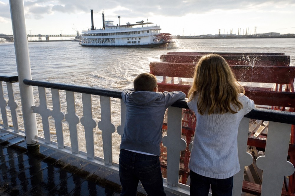 Two children leaning on the white railing of a riverboat deck watching the paddlewheel churn brown river water