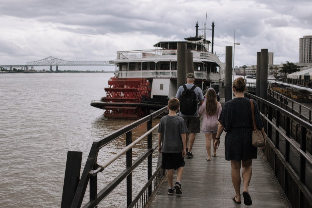 Family of four walking down a wooden wharf ramp toward a moored white riverboat with a red paddlewheel