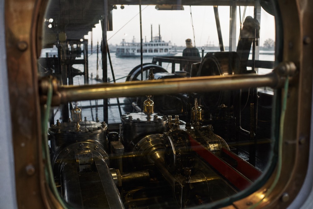 View through a brass-railed window into a riverboat engine room showing polished steam machinery in motion