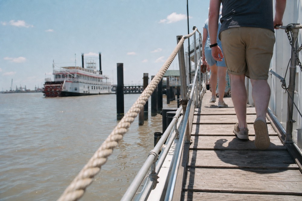 A wooden riverboat gangway with a rope handrail leading up to a white boarding deck, passengers' legs and casual shoes visible mid-step