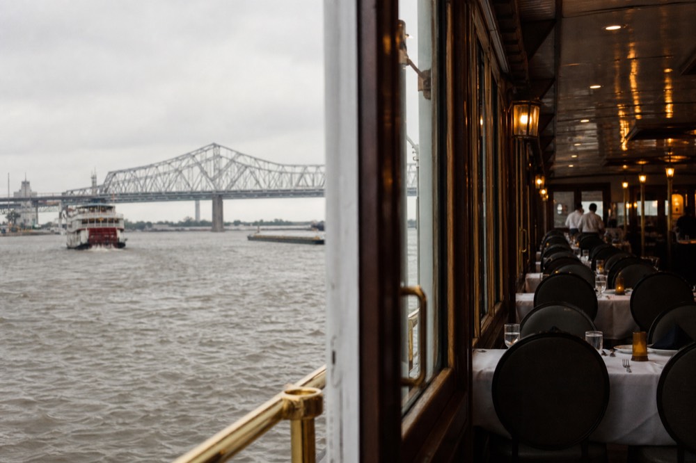 Evening riverboat dining room with white linen tablecloths, brass rails and warm lamps, seen from the doorway with empty chairs