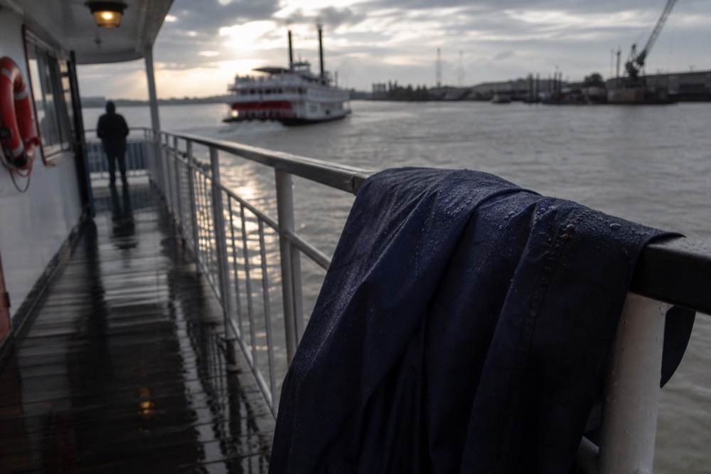 Rain beading on the shoulder of a folded waterproof shell hung over a riverboat deck rail, wet planking and grey river beyond