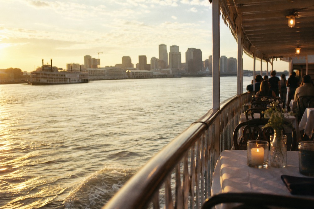 Golden late-afternoon light on a riverboat's upper deck railing with the curving New Orleans skyline in the distance and the paddlewheel wash below